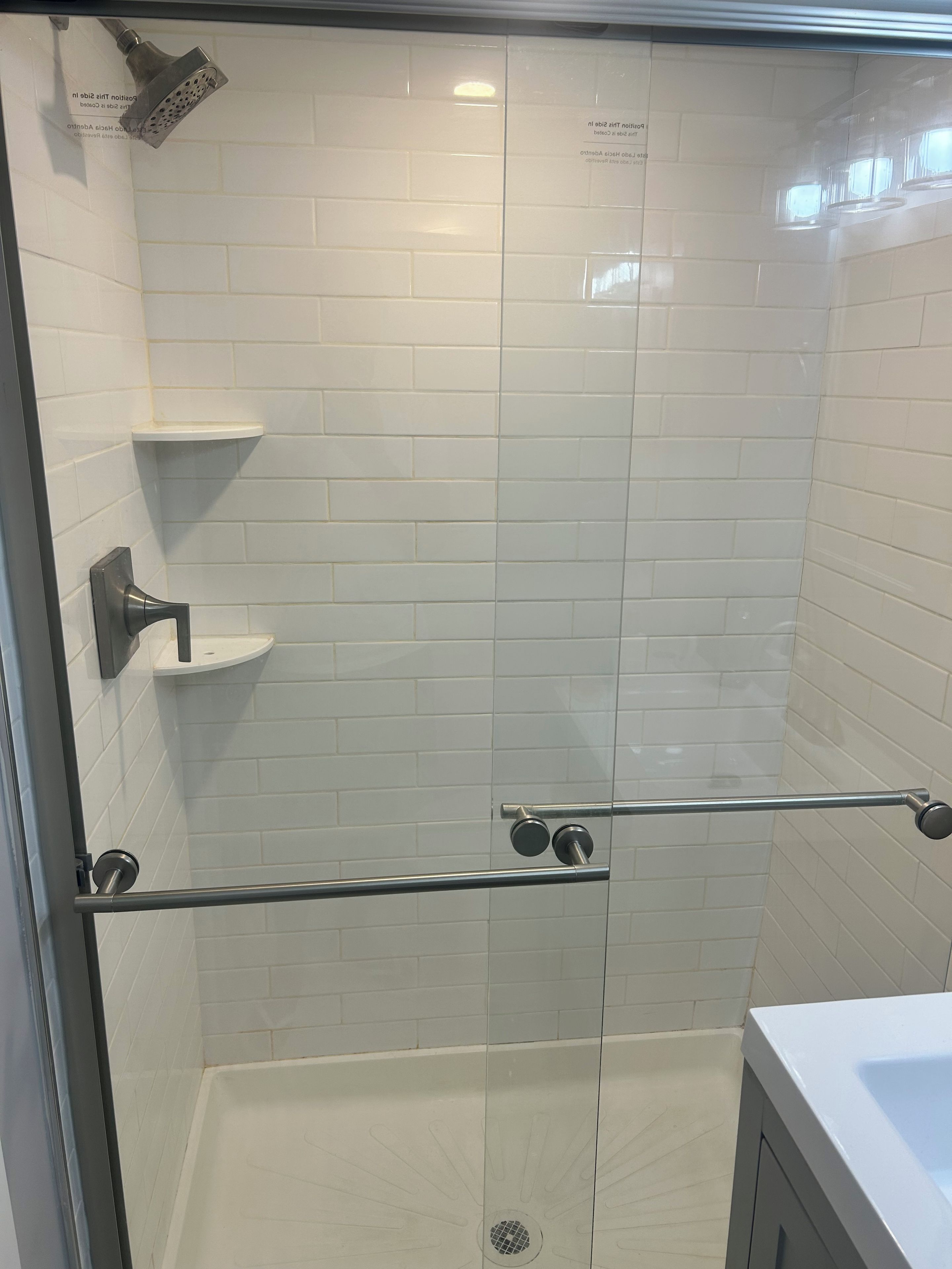 Ducha con mampara de vidrio, azulejos blancos, barra de apoyo metálica y estante.