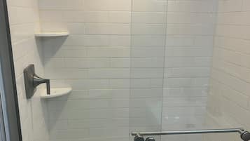 Ducha con mampara de vidrio, azulejos blancos, barra de apoyo metálica y estante.