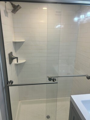 Ducha con mampara de vidrio, azulejos blancos, barra de apoyo metálica y estante.