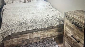 Un dormitorio con estructura de cama de madera, colcha de encaje, alfombra estampada y cómoda de madera con cajones.