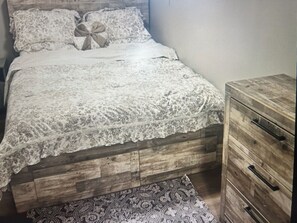 Un dormitorio con estructura de cama de madera, colcha de encaje, alfombra estampada y cómoda de madera con cajones.