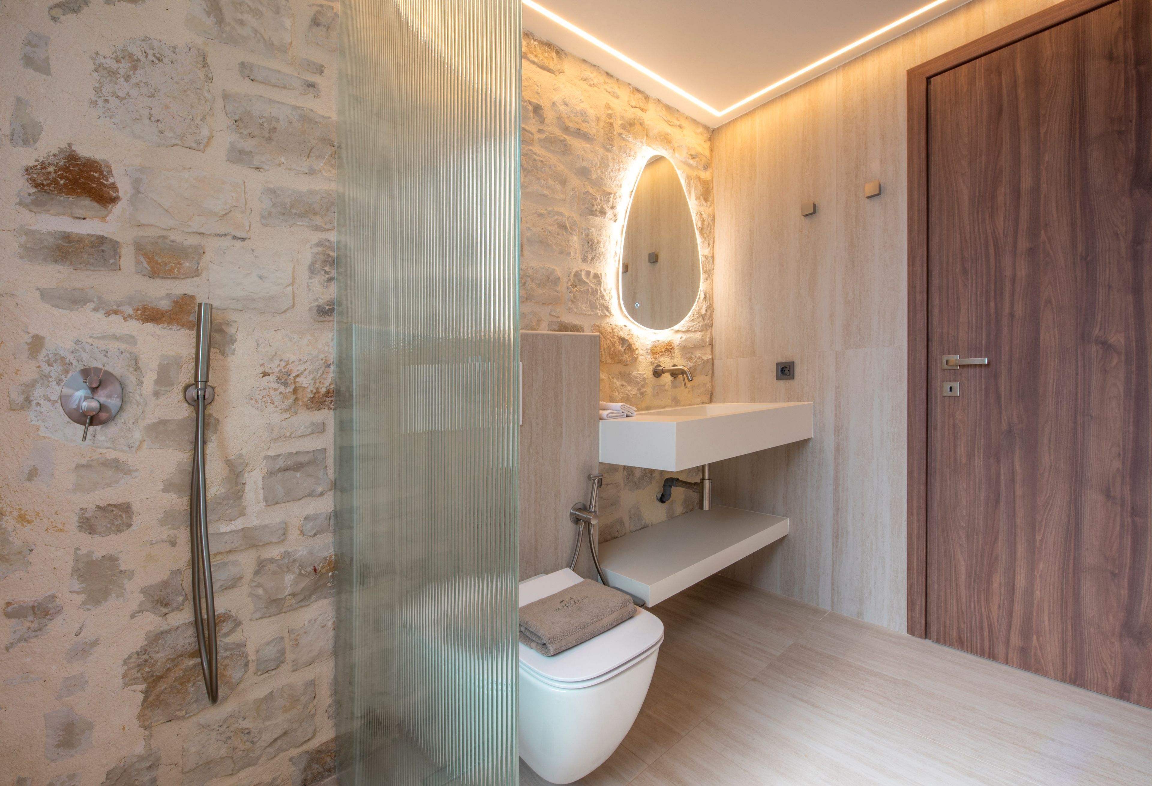 Un baño moderno con pared de piedra, cabina de ducha de vidrio, puerta de madera y lavamanos blanco.