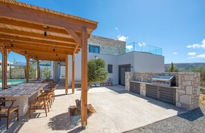 An outdoor patio with a wooden pergola, seating area, and a stone barbecue station. - Black Olive Villas (Pigi)