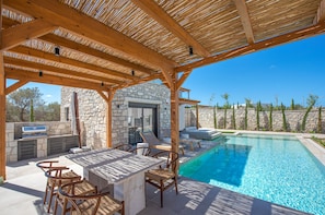 Un área de piscina al aire libre con un edificio de piedra, una pérgola de madera y mobiliario.