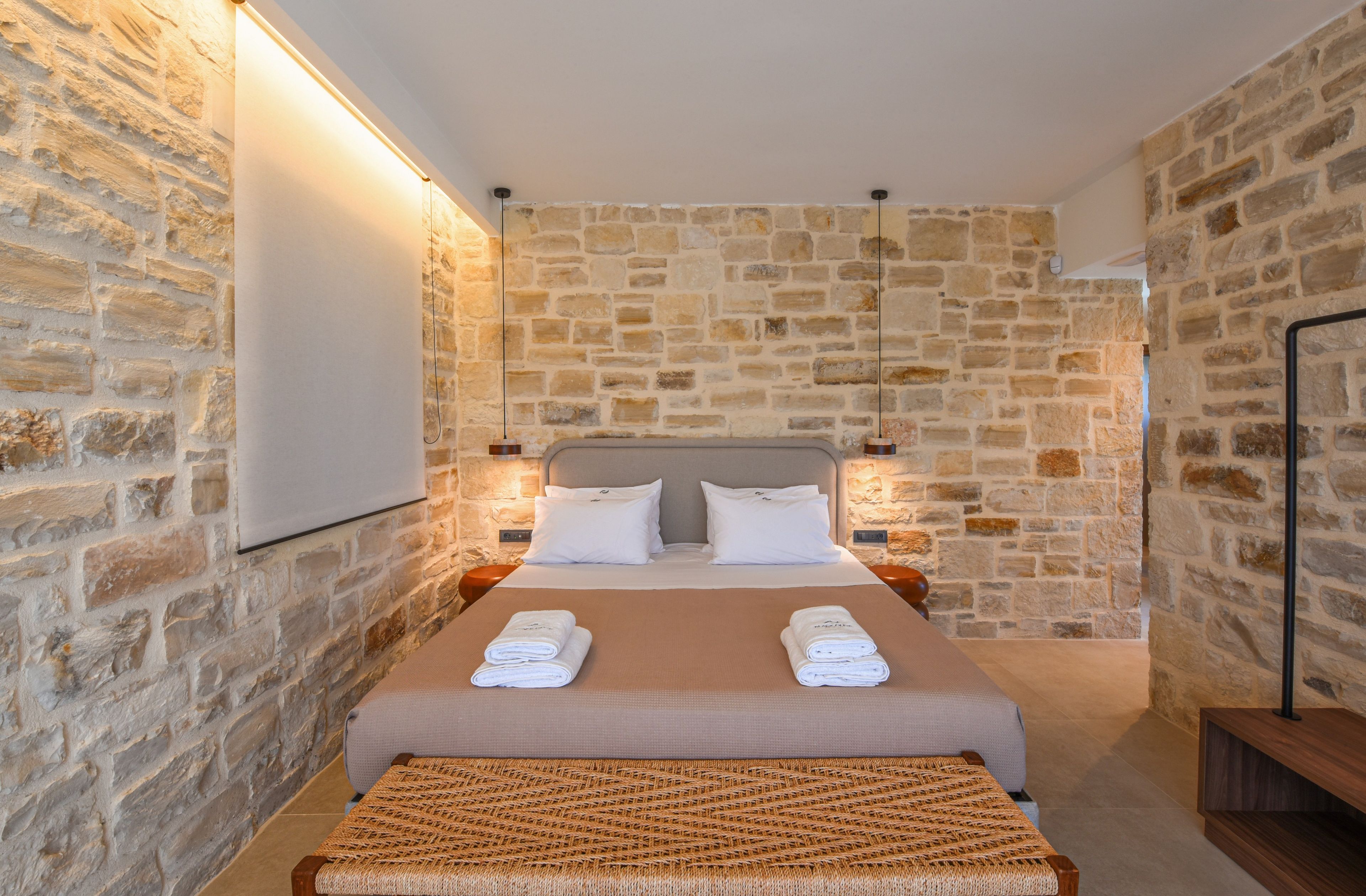 Habitación de hotel con pared de piedra, cama con sábanas blancas y un banco tejido.