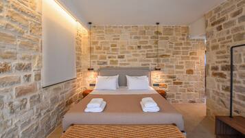 Habitación de hotel con pared de piedra, cama con sábanas blancas y un banco tejido.