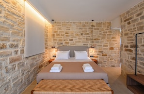 A hotel room with a stone wall, a bed with white linens, and a woven bench. - Black Olive Villas (Pigi)