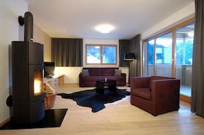 A living room with a wood-burning stove, a sofa, a chair, and a large window with a view of a snowy landscape. - Chalet With Fireplace and Garden (Gaschurn)