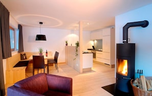 A modern living room with a sofa, a dining area with chairs and a table, a kitchen with cabinets and a stove, and a large window with curtains. - Chalet With Fireplace and Garden (Gaschurn)