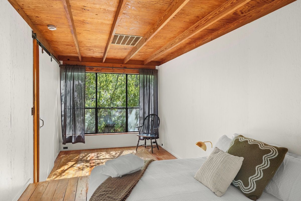 Une pièce avec un plafond en bois, un lit avec des oreillers, une chaise et une fenêtre avec des rideaux.