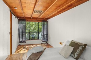 A room with a wooden ceiling, a bed with pillows, a chair, and a window with curtains.