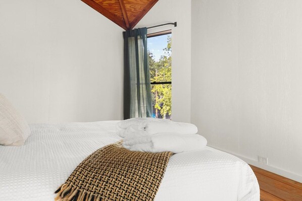 A neatly made bed with white linens and a woven throw blanket, a window with a view of trees, and a wooden ceiling beam.