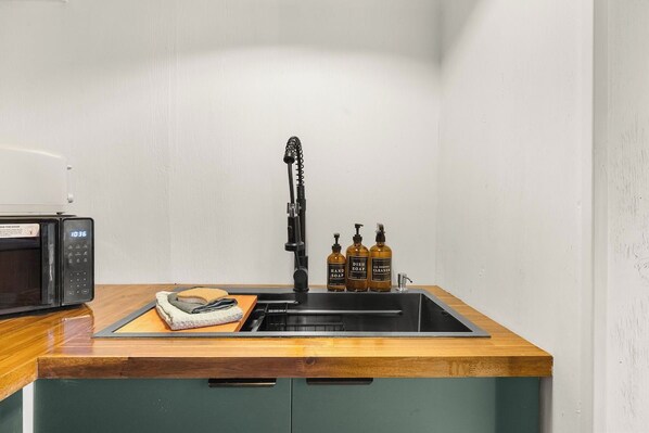 A kitchen with a wooden countertop, a black sink, a microwave, and three bottles of hand soap.