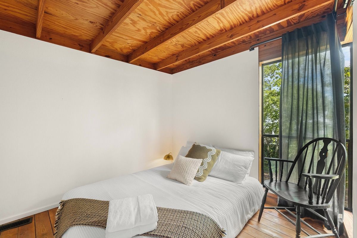 Une chambre avec un plafond en bois, un lit avec une literie blanche, une chaise et une fenêtre avec des rideaux.