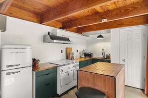 Cocina compacta con un refrigerador blanco, estufa y microondas. Se observa un techo de madera y una puerta blanca al fondo.