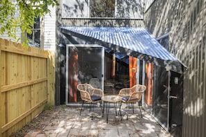 An outdoor patio with two wicker chairs and a table, a wooden fence, and a building with a corrugated metal roof.