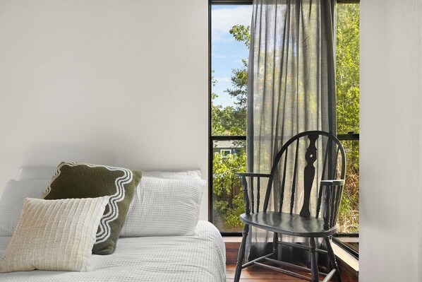 Un dormitorio con cama, almohadas, una silla y una ventana con cortinas.