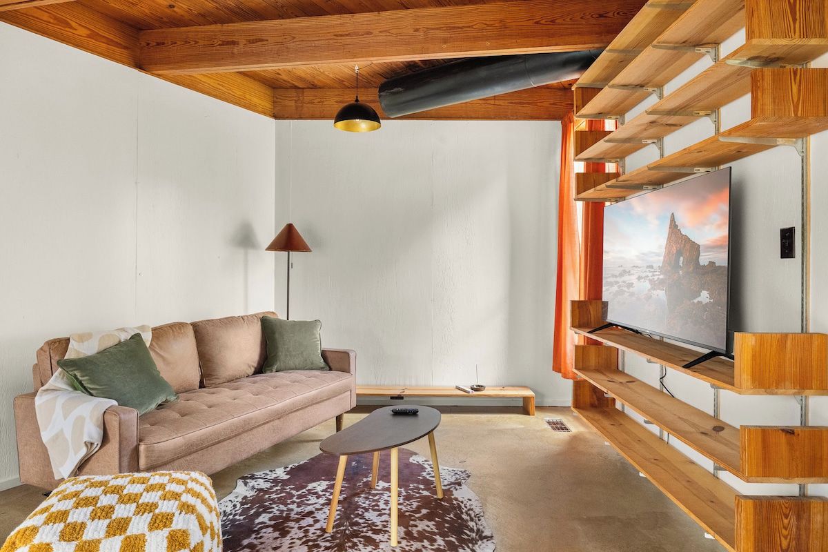 Un salon avec un canapé, une table basse, un téléviseur posé sur une étagère en bois et un luminaire suspendu.