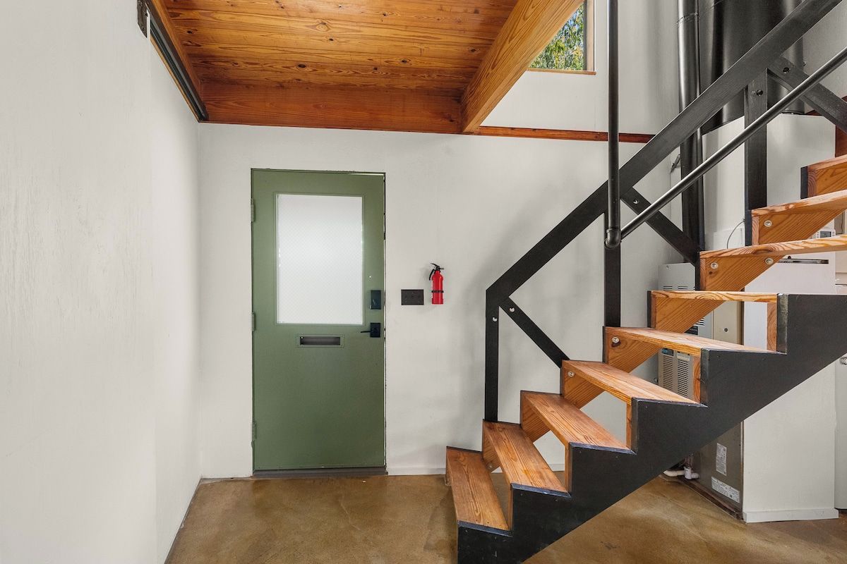 Un escalier avec des marches en bois et une rampe en métal noir, menant à une pièce dotée d’une porte verte et d’un extincteur fixé au mur.