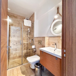 A modern bathroom with a glass shower enclosure, a toilet, a sink with a mirror, and a trash can. - A.emreli Suite Hotel (Ayvalik)