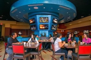 Un restaurante con varias pantallas mostrando contenido deportivo, comensales sentados en mesas y un techo azul característico.
