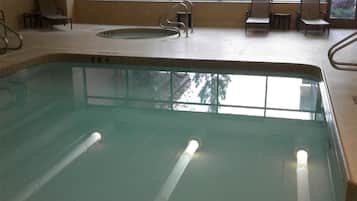 An indoor swimming pool with a clear view of the outside through large windows, a seating area with chairs, and a tiled floor.
