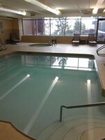 An indoor swimming pool with a clear view of the outside through large windows, a seating area with chairs, and a tiled floor.