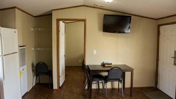 A compact room with a kitchenette, a dining table with chairs, a television mounted on the wall, and a door leading to another room.