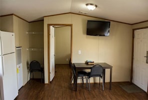 Quarto compacto com cozinha americana, mesa de jantar com cadeiras, televisão na parede e porta para outro cômodo.