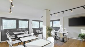 A modern conference room with white tables and chairs, large windows, a flat-screen TV, and a ceiling track lighting system. - Ibis Budget Berlin Genshagen (Ludwigsfelde)