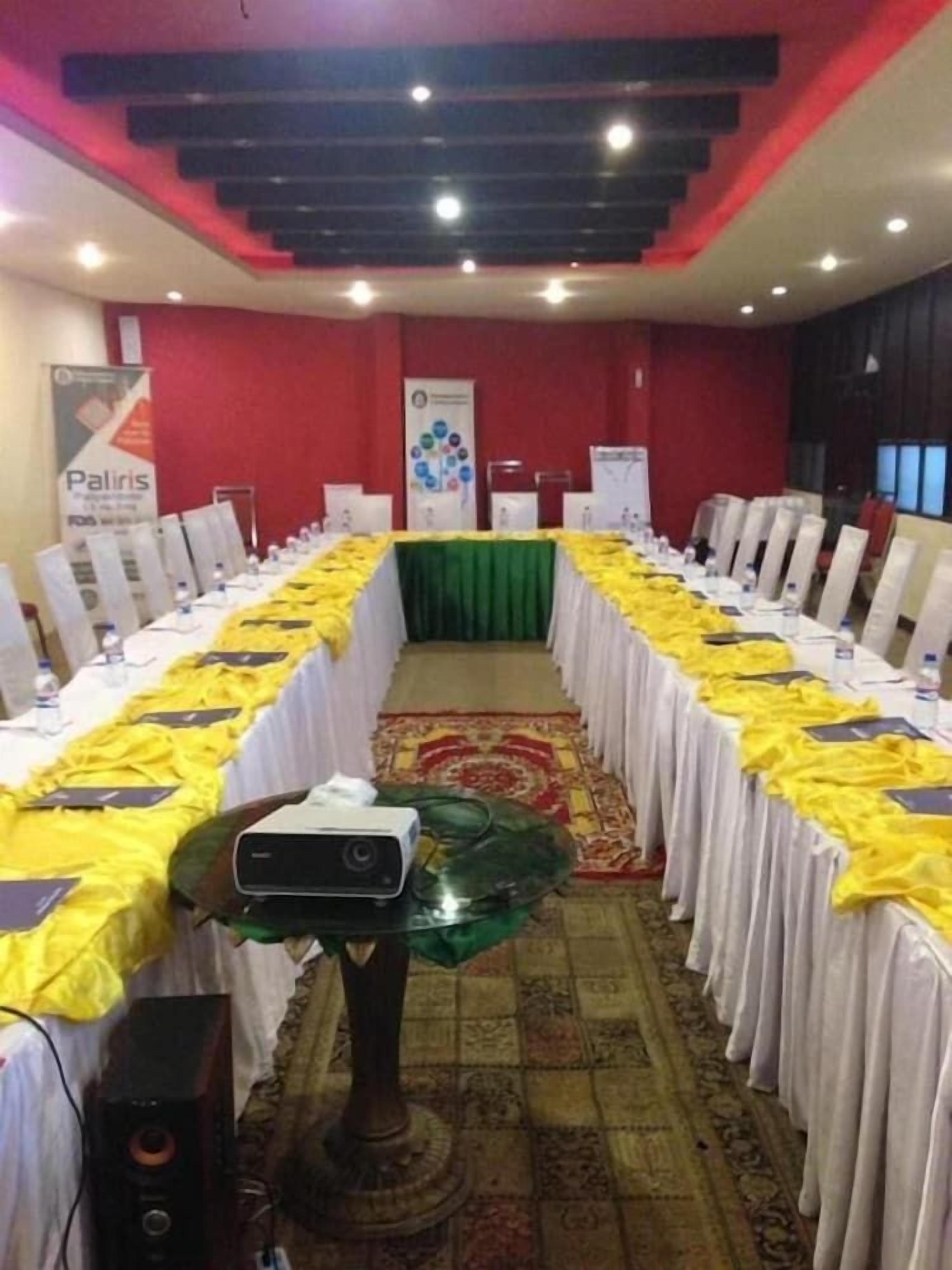 Un salón de conferencias con una larga mesa dispuesta para una reunión, un proyector sobre un soporte y un orador en primer plano.