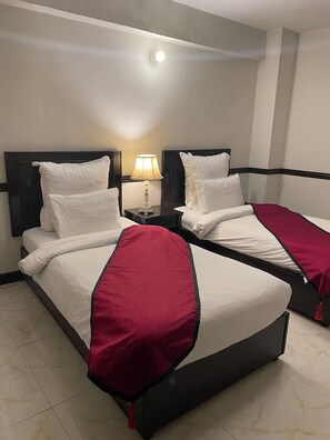 Habitación de hotel con dos camas, cada una con ropa blanca y detalles en rojo. Se ve una mesita de noche con una lámpara entre las camas.
