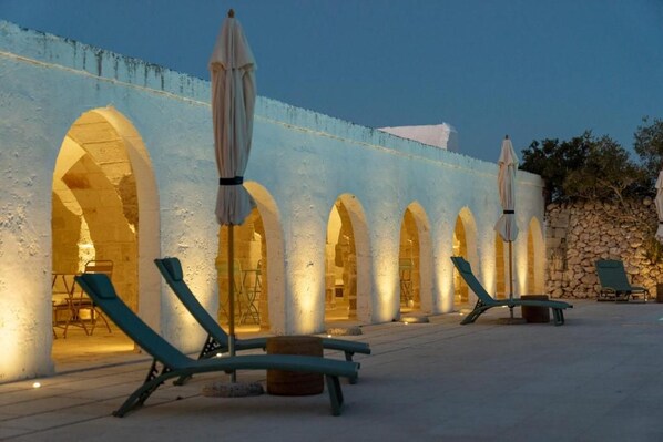 A series of outdoor lounge chairs and umbrellas aligned along a wall with arched openings.