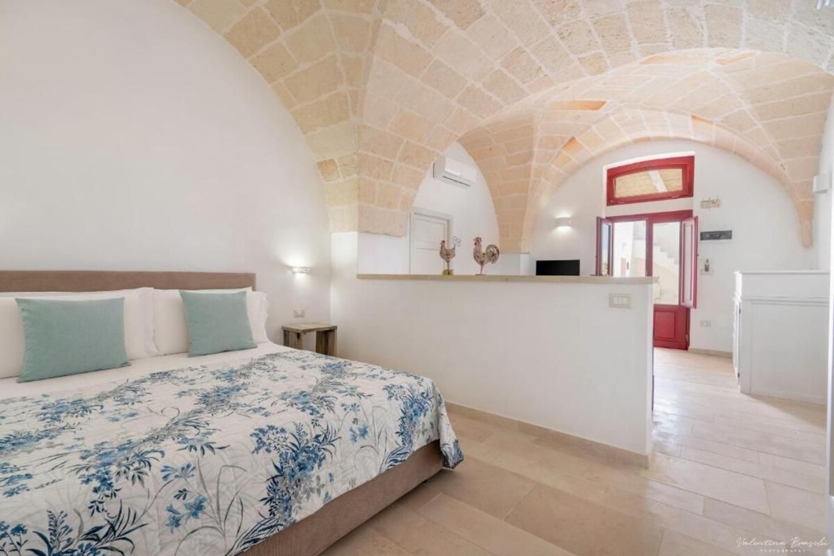 Une chambre comprenant un lit, une petite table de chevet, un banc intégré et un coin cuisine avec un comptoir et une porte.