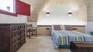 Una camera da letto con soffitto a volta in pietra, un comò in legno, un letto con biancheria a righe e una finestra con persiane rosse.