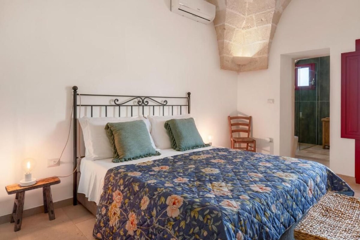 A bedroom with a bed, a bedside table with a lamp, a chair, and a red door.