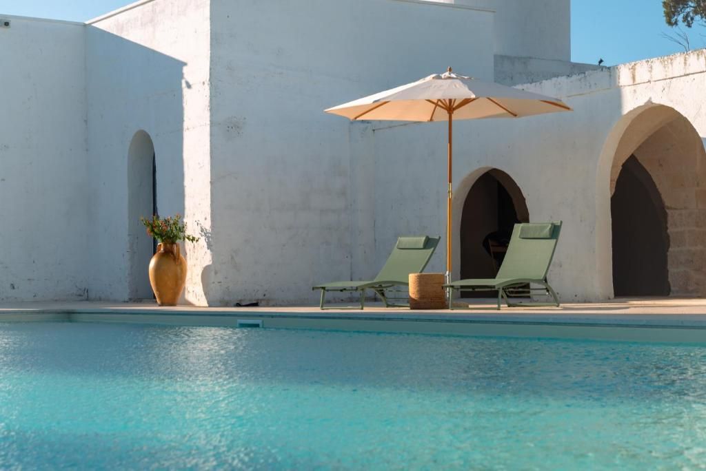 Un área de piscina con dos sillones de descanso y una sombrilla, una planta en maceta y un edificio con arcos.