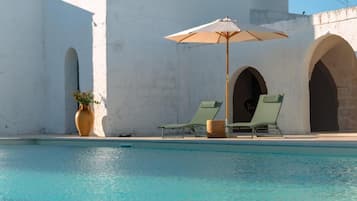 Un área de piscina con dos sillones de descanso y una sombrilla, una planta en maceta y un edificio con arcos.