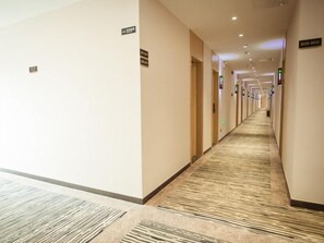 A hotel corridor with a carpeted floor, doors on the right, and signs indicating room numbers.