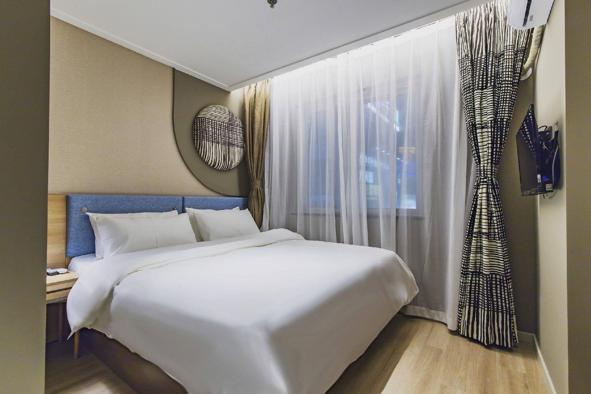 A hotel room with a bed, a bedside table, a wall-mounted TV, and a window with sheer curtains.
