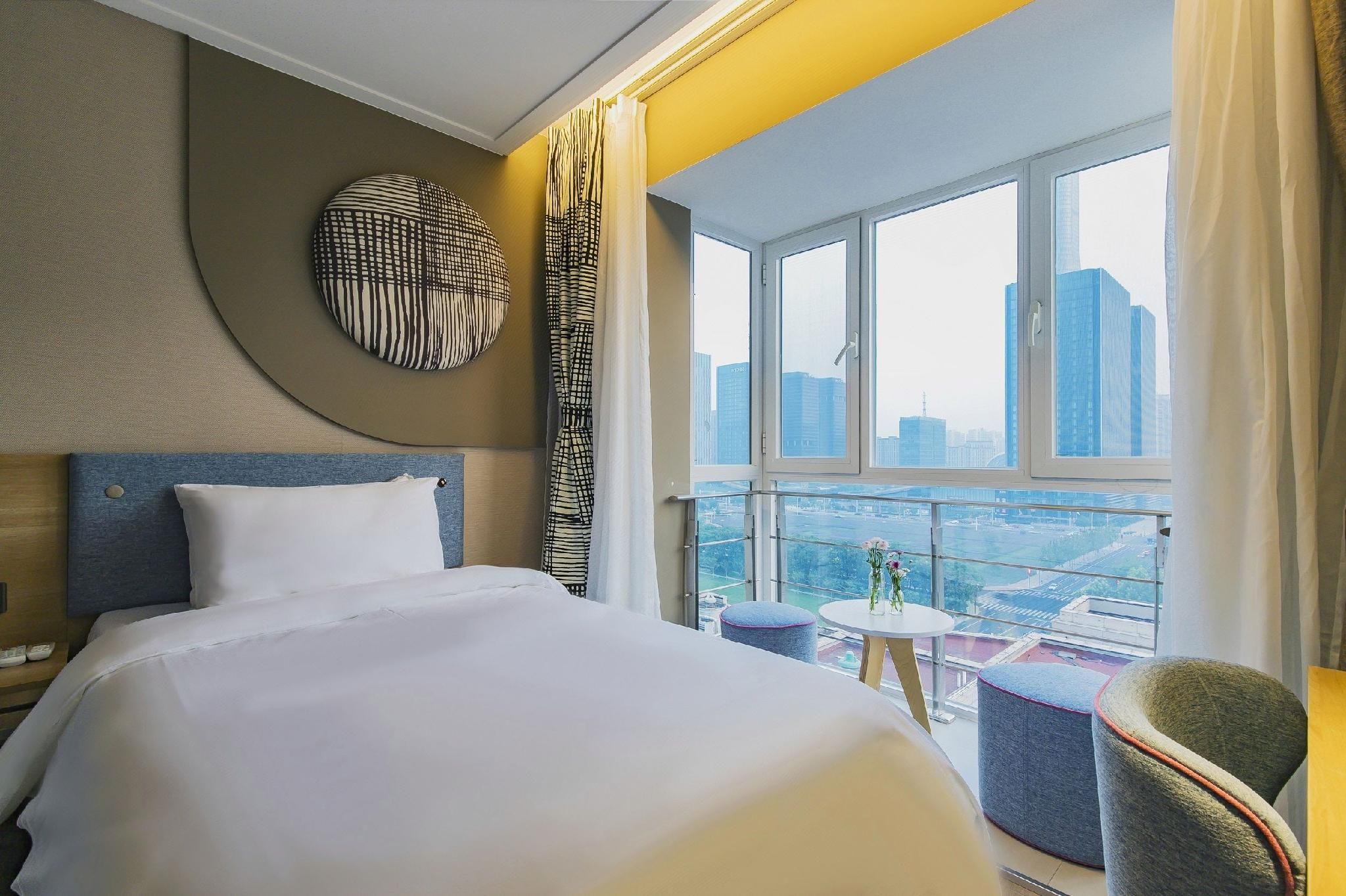 A hotel room with a large bed, a round wall decoration, a view of the cityscape through a large window, and a small table with flowers.