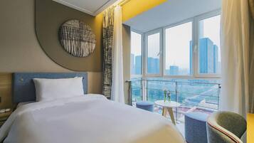 A hotel room with a large bed, a round wall decoration, a view of the cityscape through a large window, and a small table with flowers.