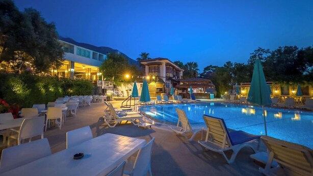 A poolside area with lounge chairs and tables, a pool, and buildings in the background. - Zehra Tatil Koyu (Fethiye)