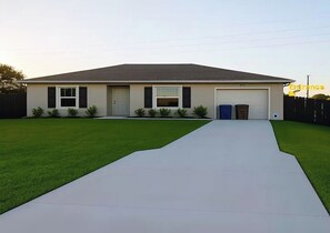 A single-story house with a garage, a front yard with artificial grass, and a driveway leading to the garage. - Private Studio Near RSW & JetBlue Park (Lehigh Acres)