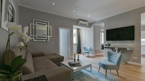 Una sala moderna con sofá, mesa de centro y televisor. Se ve una puerta que da a otra habitación y una ventana con cortinas.