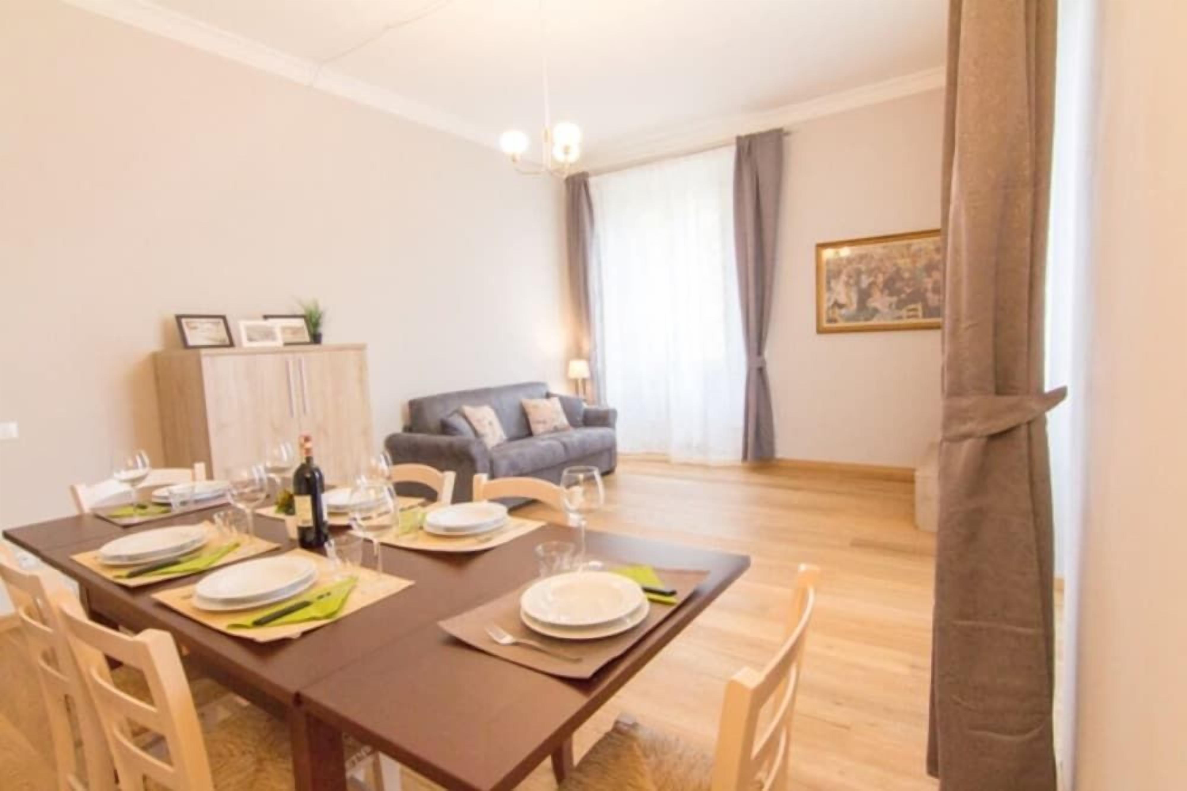 A dining area with a wooden table set for four, a sofa in the background, and a window with curtains.