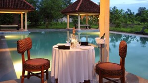 A dining table with two wicker chairs, a candle, and a bottle, set by a poolside with a gazebo in the background. - Tuli Tiger Corridor (Barghat)