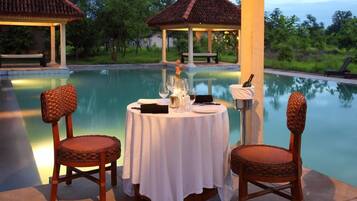 A dining table with two wicker chairs, a candle, and a bottle, set by a poolside with a gazebo in the background.