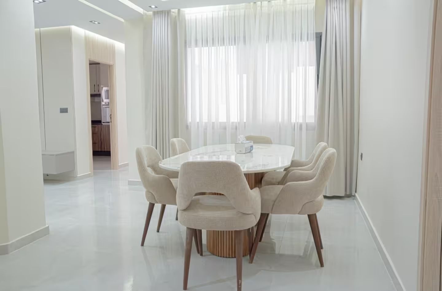 Un espace repas moderne avec une table ronde et six chaises, une cuisine en arrière-plan et des rideaux légers aux fenêtres.