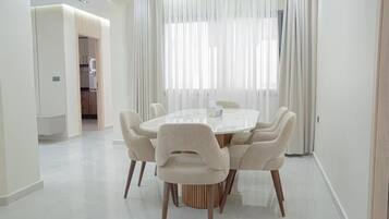 Un espace repas moderne avec une table ronde et six chaises, une cuisine en arrière-plan et des rideaux légers aux fenêtres.